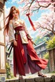 A woman in traditional attire stands outdoors with cherry blossoms and traditional buildings in the background.